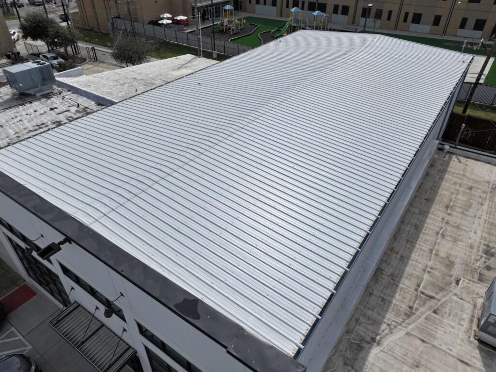 An aerial photo shows the large, flat, corrugated metal roof of a building. The roof is light gray and has numerous parallel ridges. The building's walls are white with black trim. In the background, there is a playground and another building, and a few cars are visible in a parking lot. The ground is a light gray concrete.