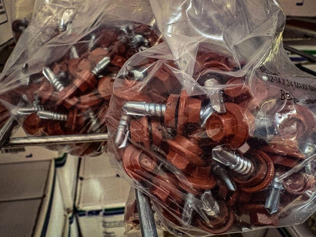 A close-up, high-angle photo of two clear plastic bags filled with brown-colored roofing screws. The screws have a silver-colored tip and a hexagonal head with an attached washer. The bags are tied at the top and are hanging from a metal rack. The background is a blurry warehouse or hardware store setting.