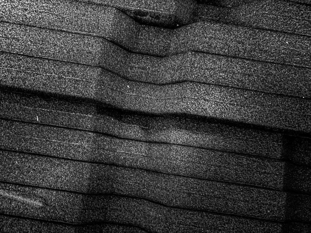 A black and white, high-contrast close-up photo of a black, textured, and ridged material. The ridges run horizontally across the image, and a slight curve is visible at the top. The texture is grainy, and a prominent diagonal shadow divides the image.