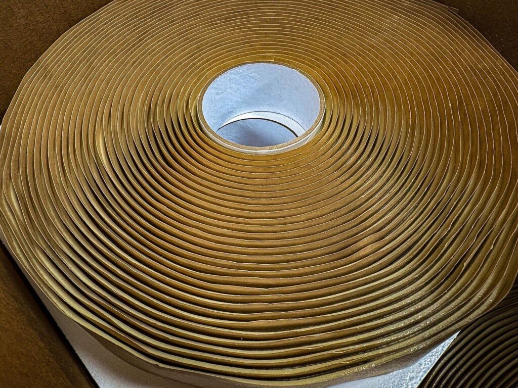 A close-up, high-angle photo of a large roll of brown or tan-colored foam sealant tape. The roll has a white inner core and is tightly wound, showing the multiple layers. The roll is sitting inside an open cardboard box, and another roll is partially visible in the bottom right corner.