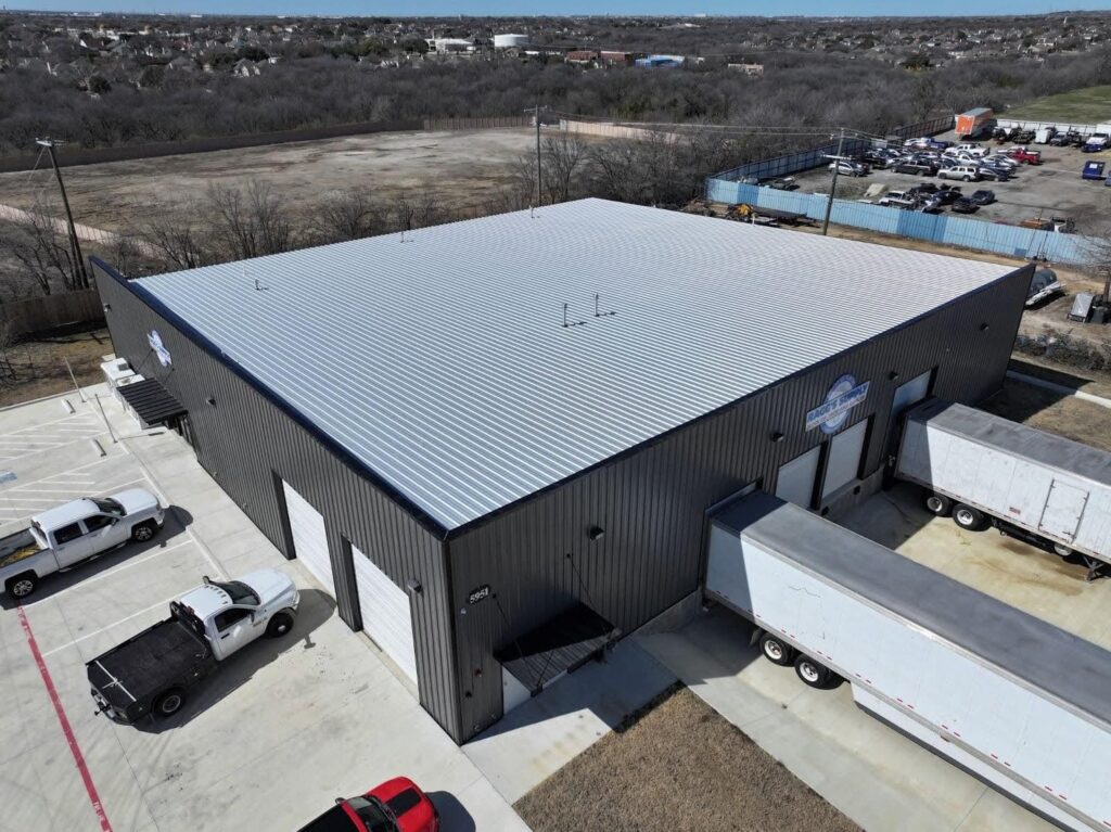 An aerial photo shows a modern, black metal industrial building with a large, shiny silver metal roof. Two large white tractor-trailer trucks are backed up to loading docks on the right side of the building. Two white pickup trucks are parked near the front of the building on the left. The building is surrounded by paved ground, and the background is a mix of a large, flat field and a forest of bare trees under a clear blue sky.
