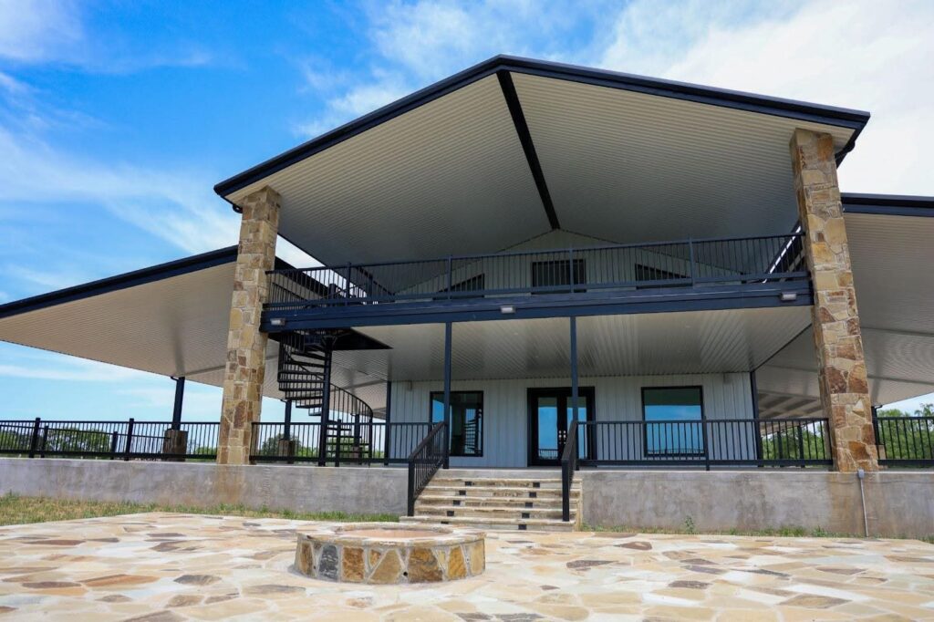 A professional photo of a large, modern home with a metal exterior and a multi-tiered design. The building has a prominent front porch and a second-story balcony, both with black metal railings. A spiral staircase connects the two levels on the left. The roof is a black metal overhang, and the walls are a light-colored metal siding. Two large pillars with a stone facade support the roof. A patio area with stone pavers and a matching stone fire pit is in the foreground. The sky is a bright blue with scattered clouds.