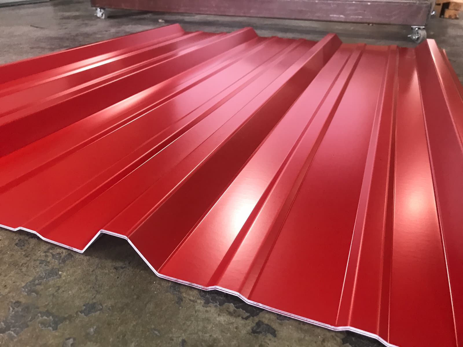 A low-angle photo of a large, single sheet of bright red metal roofing. The sheet has a corrugated, ridged pattern running horizontally from left to right, creating a sense of depth and reflection. The sheet is sitting on a gray, polished concrete floor.