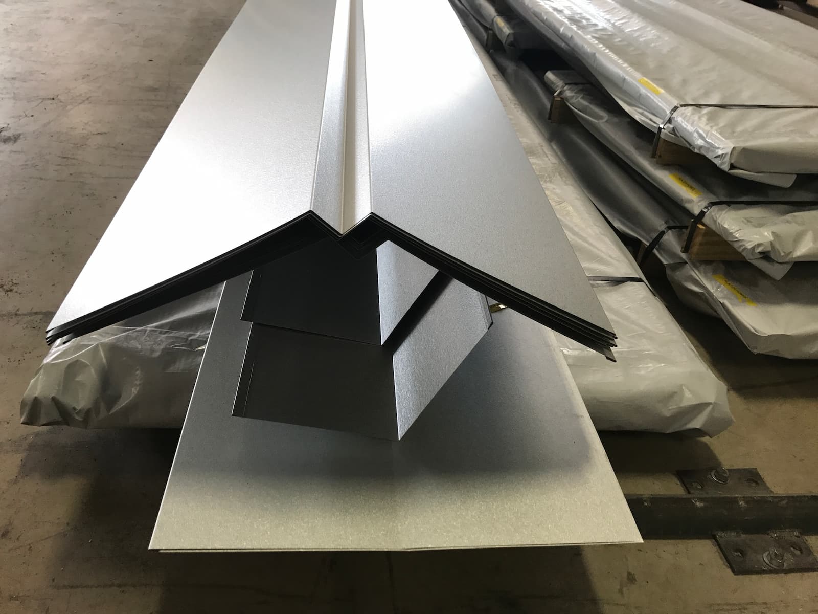 A low-angle photo of a large stack of light gray, folded metal trim pieces, likely for roofing or siding. The pieces are stacked on a wooden pallet and are protected by a white plastic sheet. The trim is folded into a sharp V-shape, and the stack stretches away from the viewer. The background is a blurry warehouse with other stacks of metal sheets.