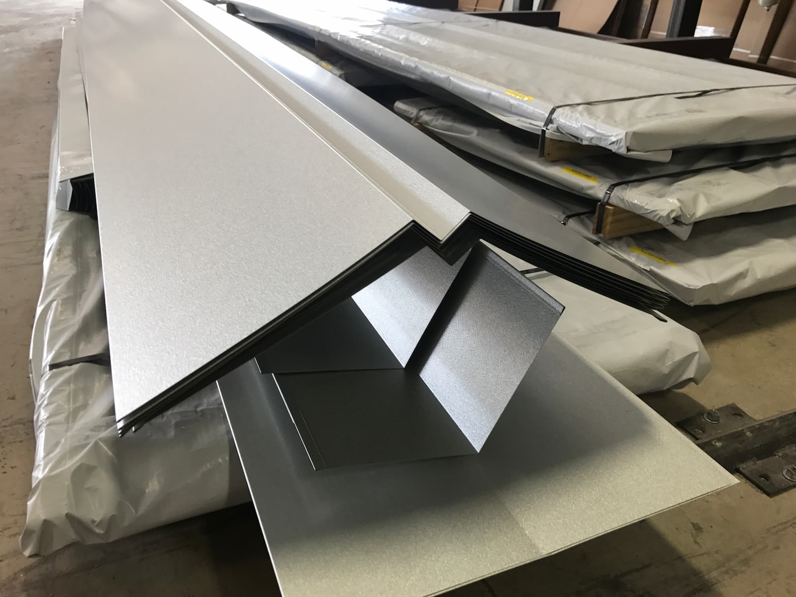 A professional photo of a large stack of light gray, folded metal trim pieces, likely for roofing or siding. The pieces are stacked on a wooden pallet and are protected by a white plastic sheet. The trim is folded into a sharp V-shape, and the stack stretches away from the viewer. The background is a blurry warehouse with other stacks of metal sheets.