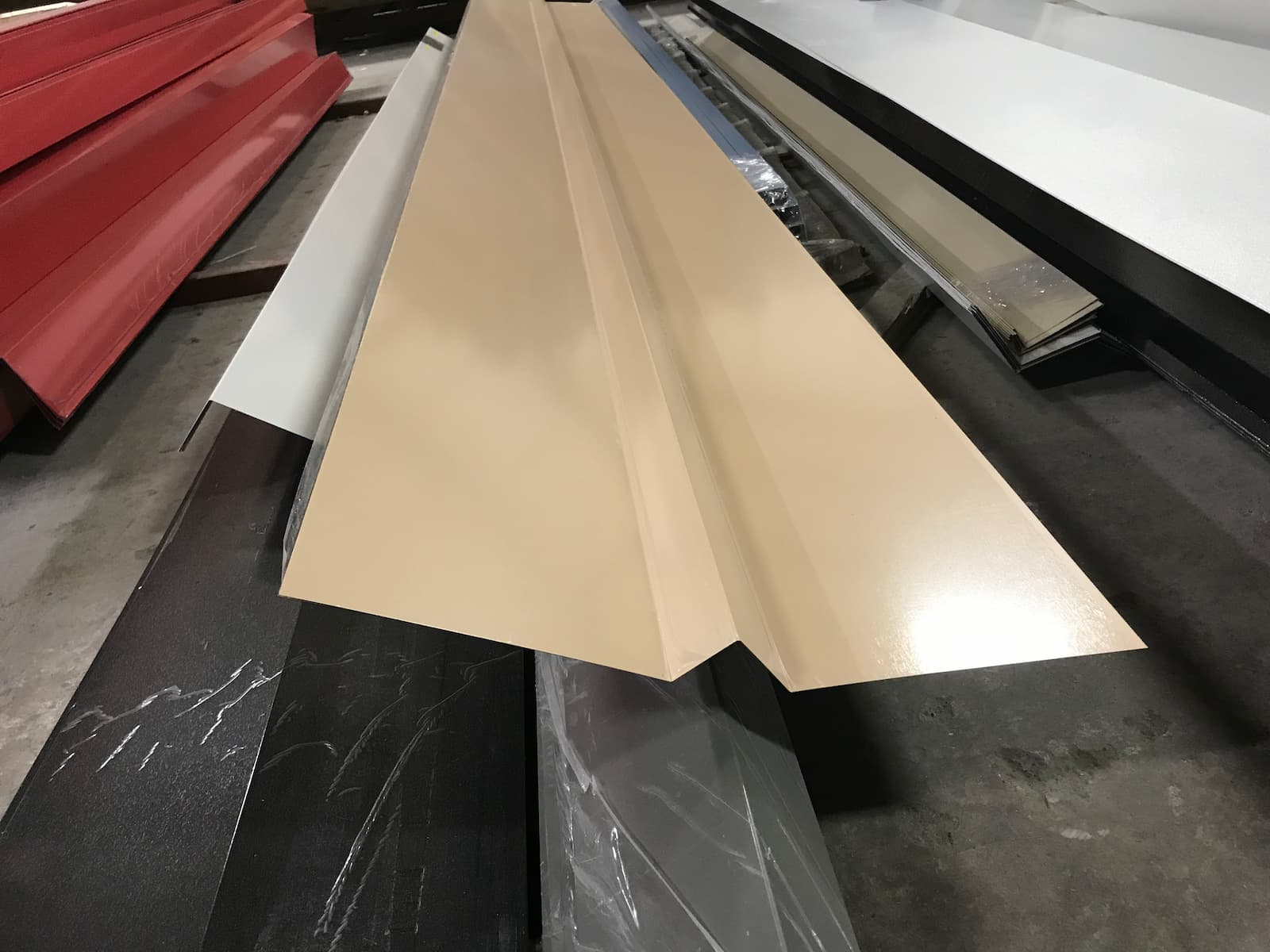 A high-angle photo of a long, folded piece of light tan or beige-colored metal trim. The trim is V-shaped and is resting on a stack of other metal pieces of different colors, including red and black marble-patterned sheets. The background is a blurry warehouse.