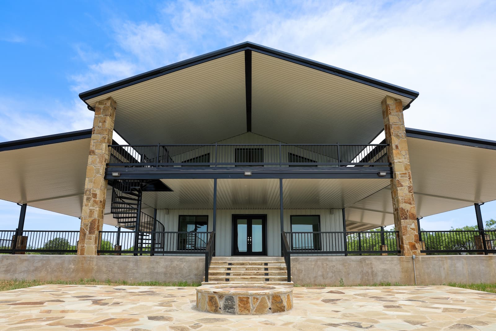 A symmetrical photo shows the front of a two-story home with a metal exterior and a large covered porch and balcony. The front has a black metal railing and a black metal spiral staircase on the left side. The roof is dark with a wide overhang and a light-colored underside. The porch is a large stone patio with a matching stone fire pit in the foreground. The sky is bright blue with scattered clouds.